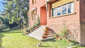 Una casa de dos pisos con una escalera que lleva a la entrada, rodeada de follaje y plantas en macetas.