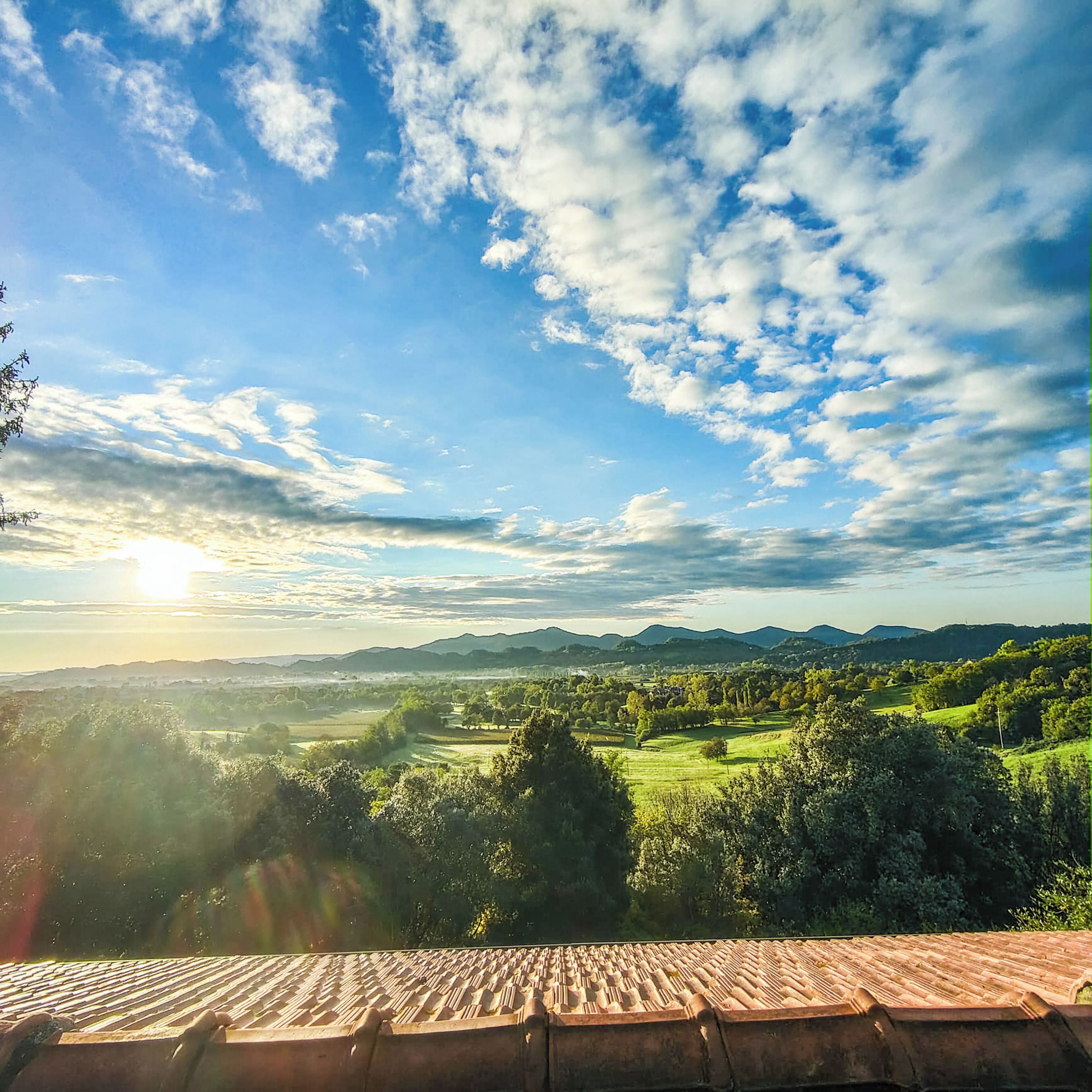 A view from a rooftop terrace with a tiled roof, overlooking a green landscape with trees and distant hills.