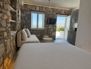 A bedroom with stone walls, a bed, a small table, a TV, and a view of the sea. - Aegean View Apartments (Ornos)