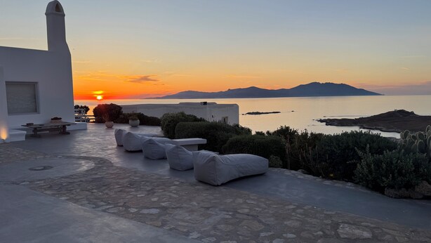 A terrace with stone flooring, white sofas, and a view of the sea at sunset. - Aegean View Apartments (Ornos)