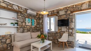 A living room with a stone wall, a sofa, a white coffee table, a TV, and a chair. - Aegean View Apartments (Ornos)