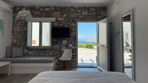 A bedroom with a stone wall, a bed, a bench, a TV, and a view of the sea. - Aegean View Apartments (Ornos)