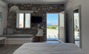 A bedroom with a stone wall, a bed, a bench, a TV, and a view of the sea. - Aegean View Apartments (Ornos)