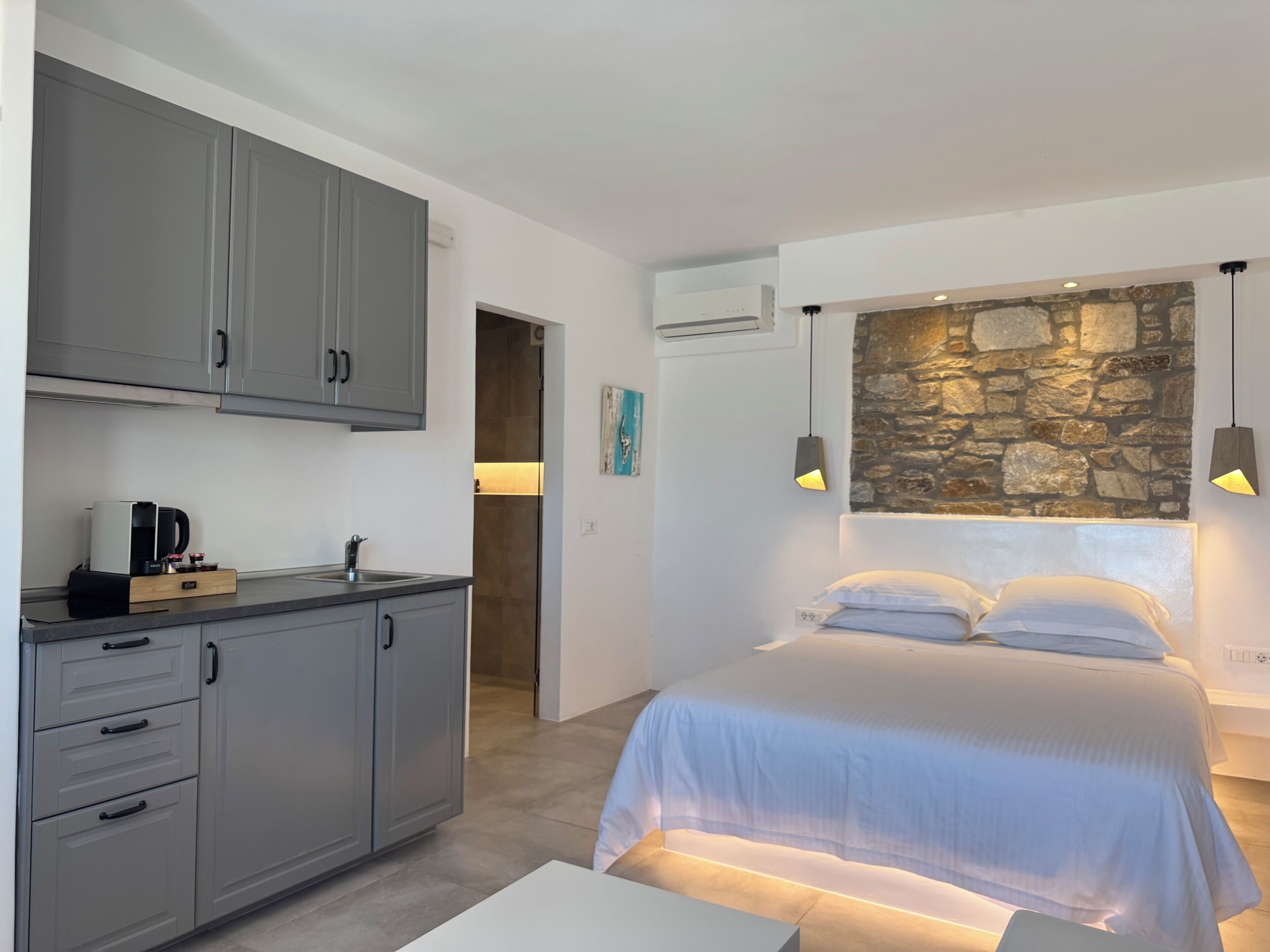 A modern bedroom with a stone accent wall, a bed with white linens, and a kitchenette with grey cabinets.