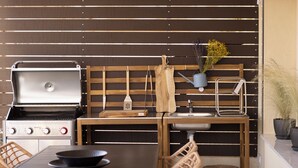 A kitchen with a grill, wooden bench, sink, and wicker chairs. - AIORA Urban Oasis - Rooftop Jacuzzi & BBQ (Thessaloniki)