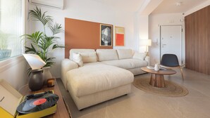 A modern hotel room with a sectional sofa, a small round table, a lamp, a potted plant, and a wall-mounted air conditioner. - AIORA Urban Oasis - Rooftop Jacuzzi & BBQ (Thessaloniki)