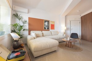 A modern hotel room with a sectional sofa, a small round table, a lamp, a potted plant, and a wall-mounted air conditioner. - AIORA Urban Oasis - Rooftop Jacuzzi & BBQ (Thessaloniki)