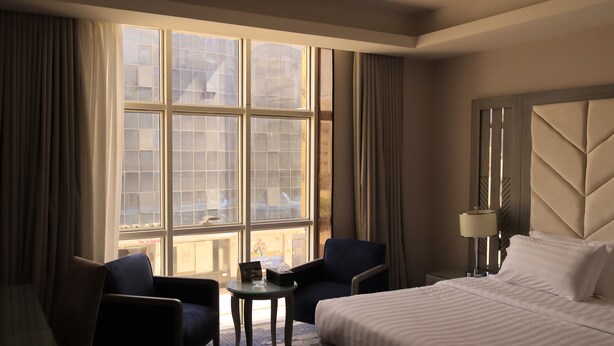 A hotel room with a large window, two armchairs, a small table, a bed, and a nightstand with a lamp. - Vercure Jeddah Alhamra Hotel (Jeddah)