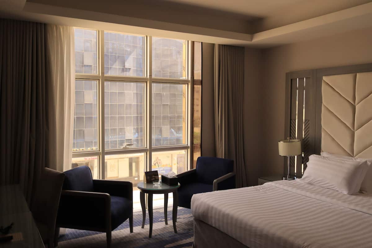 Una habitación de hotel con un ventanal grande, dos sillones, una mesita, una cama y una mesita de noche con lámpara.