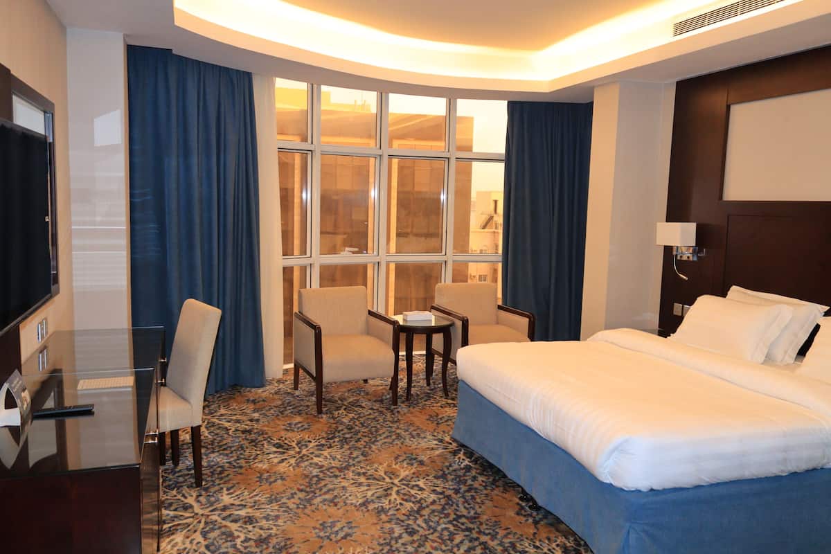Habitación de hotel con una cama, dos sillones, una mesita, un escritorio con ordenador y un amplio ventanal con cortinas.