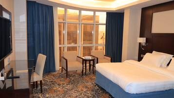Habitación de hotel con una cama, dos sillones, una mesita, un escritorio con ordenador y un amplio ventanal con cortinas.