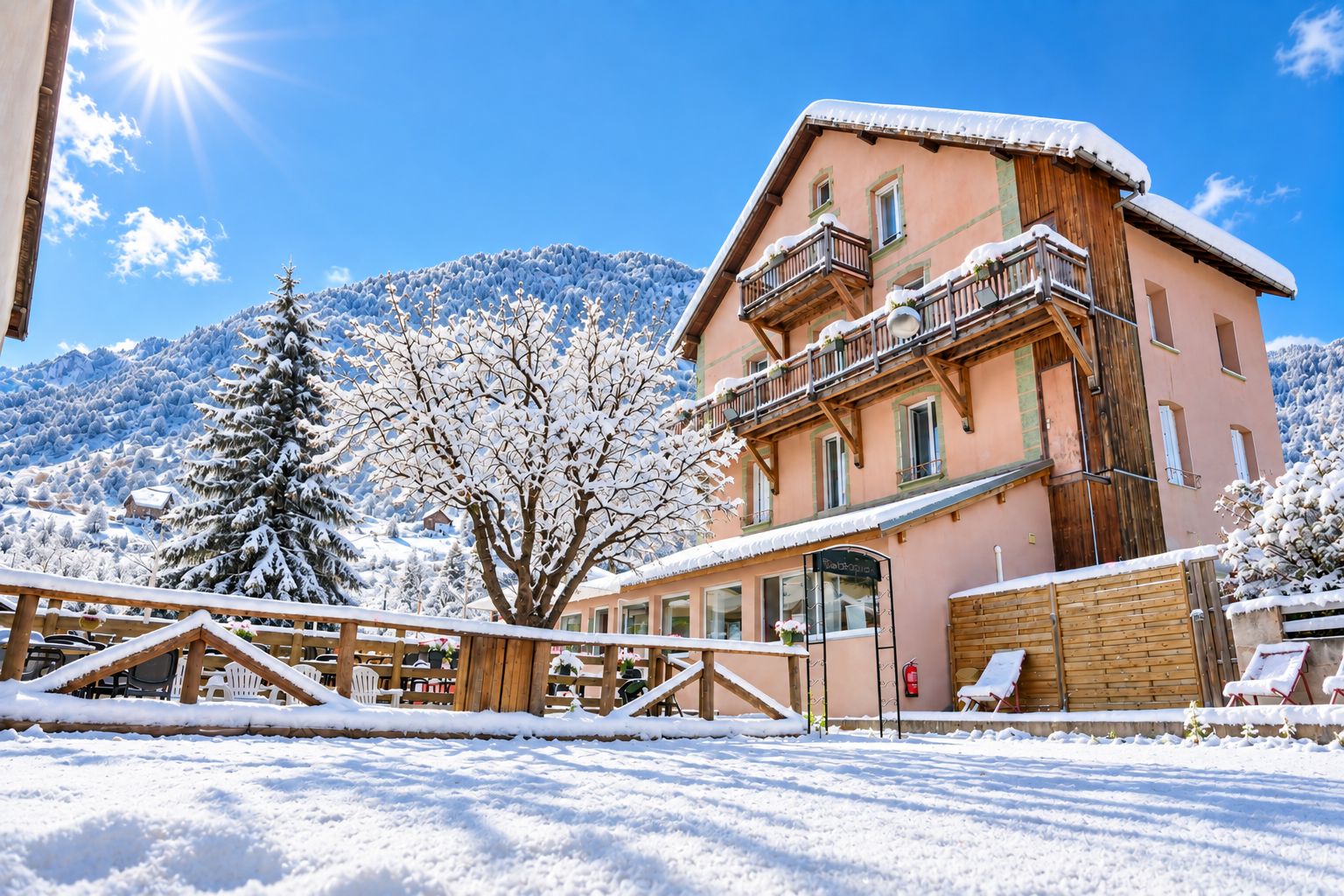 Un edificio cubierto de nieve con un balcón, rodeado de árboles nevados y montañas al fondo.