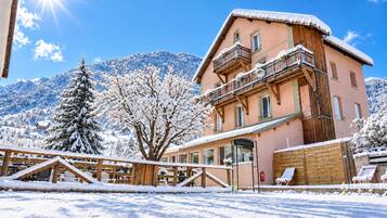 Un edificio cubierto de nieve con un balcón, rodeado de árboles nevados y montañas al fondo.