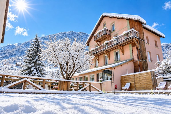 A snow-covered building with a balcony, surrounded by snow-covered trees and mountains in the background.