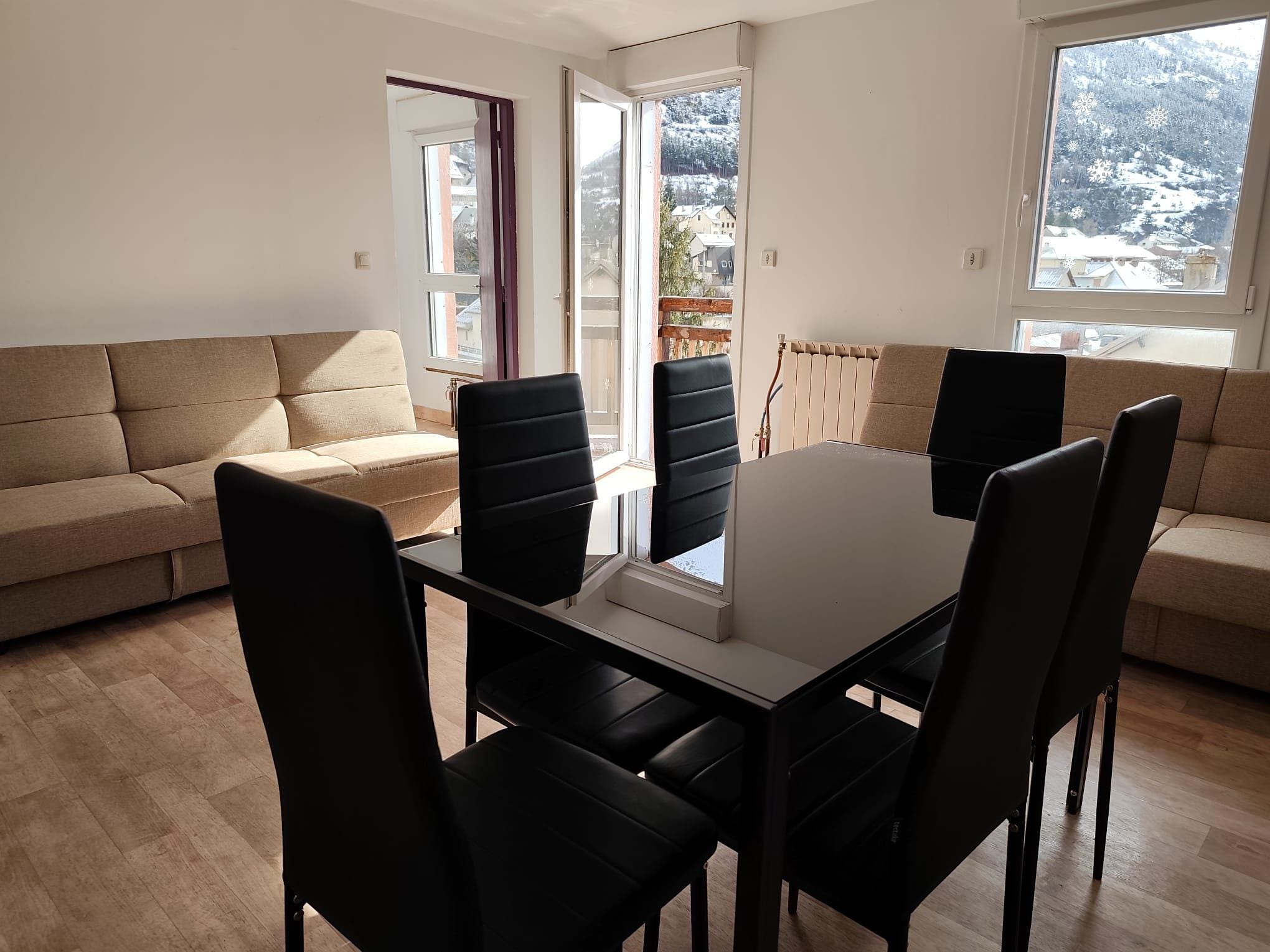 A modern living room with a beige sofa, a dining table with chairs, and large windows offering a view of a snowy landscape.