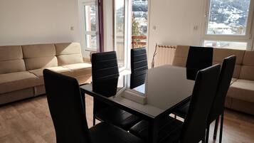 A modern living room with a beige sofa, a dining table with chairs, and large windows offering a view of a snowy landscape.