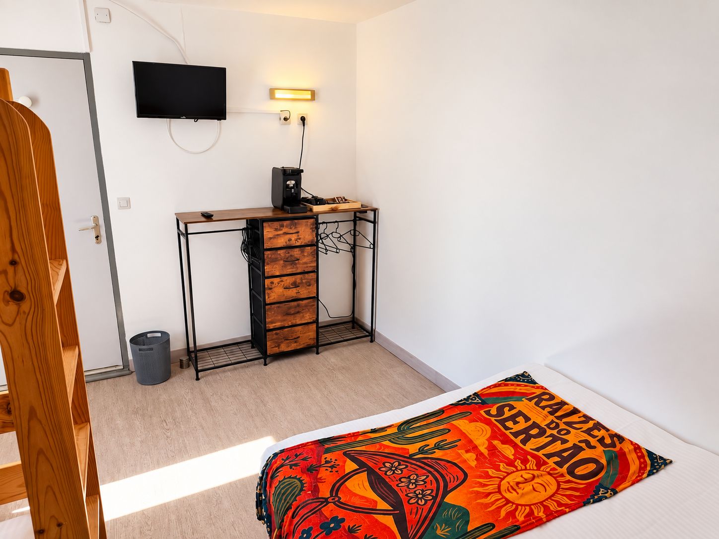 A bedroom with a bed, a desk with drawers, a television, and a wall-mounted light.