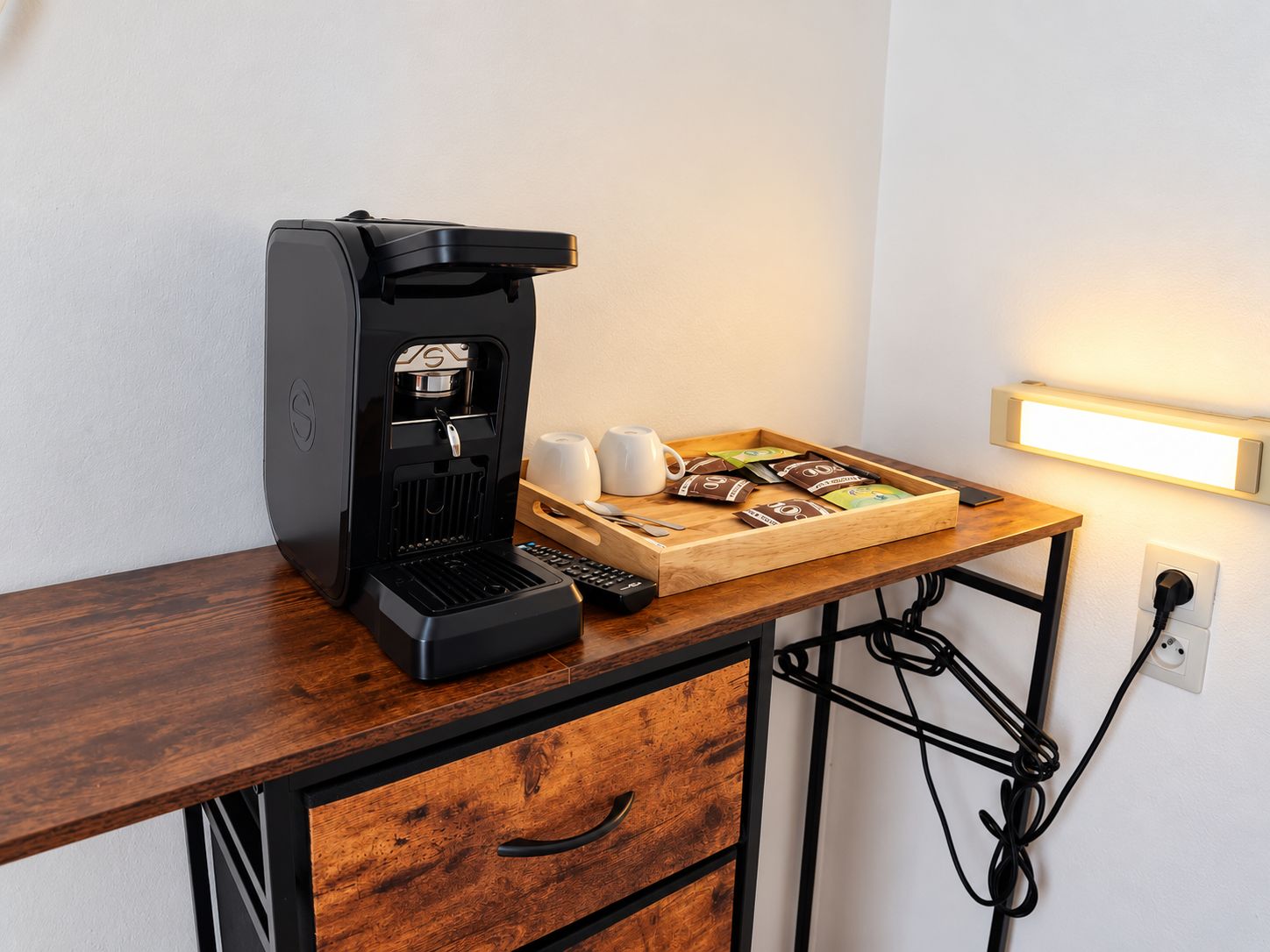 Une machine à café posée sur un bureau en bois, avec un plateau contenant des dosettes de café et des tasses, une télécommande et une prise électrique.