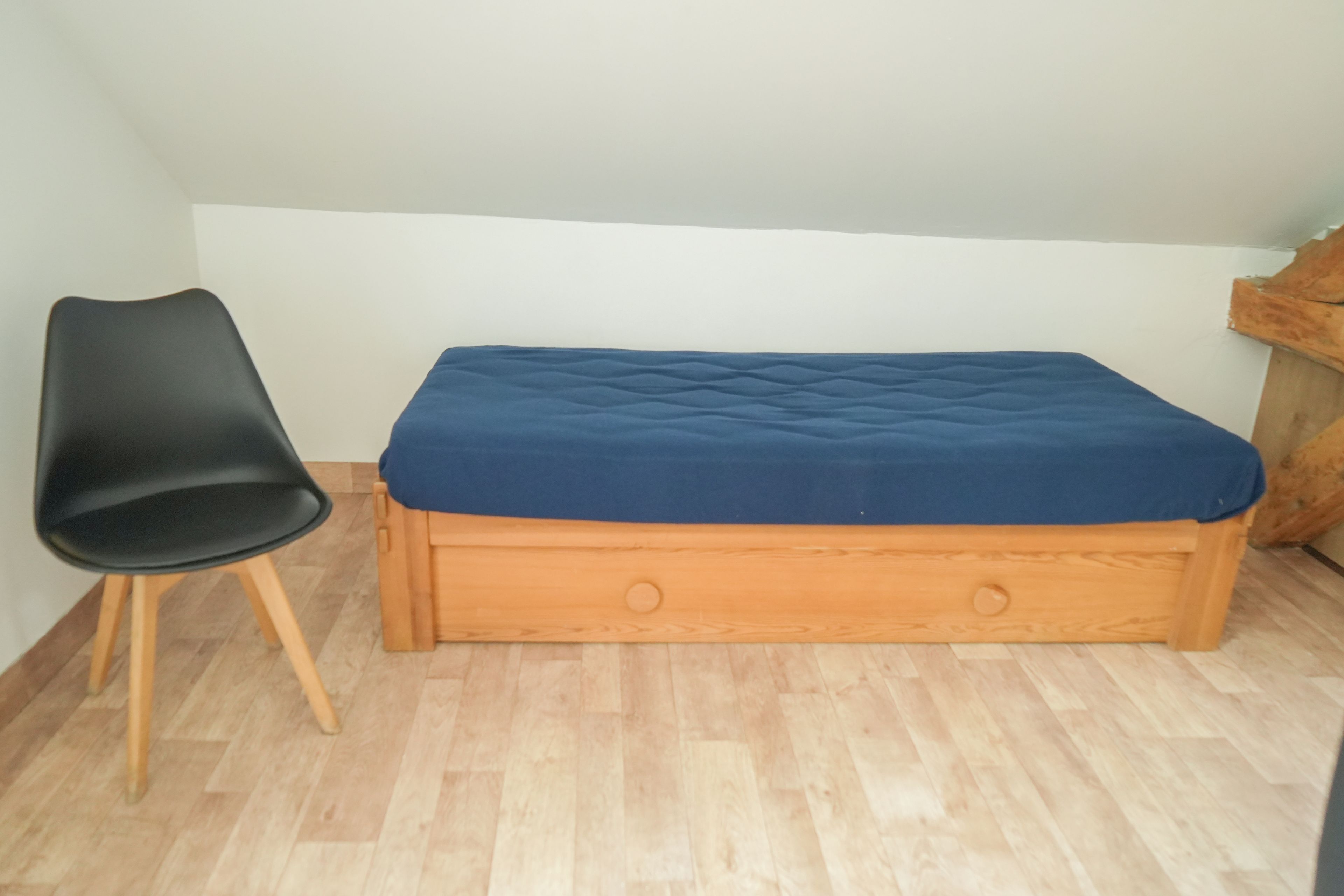 Quarto pequeno com cama de madeira, uma cadeira preta e um armário de madeira.