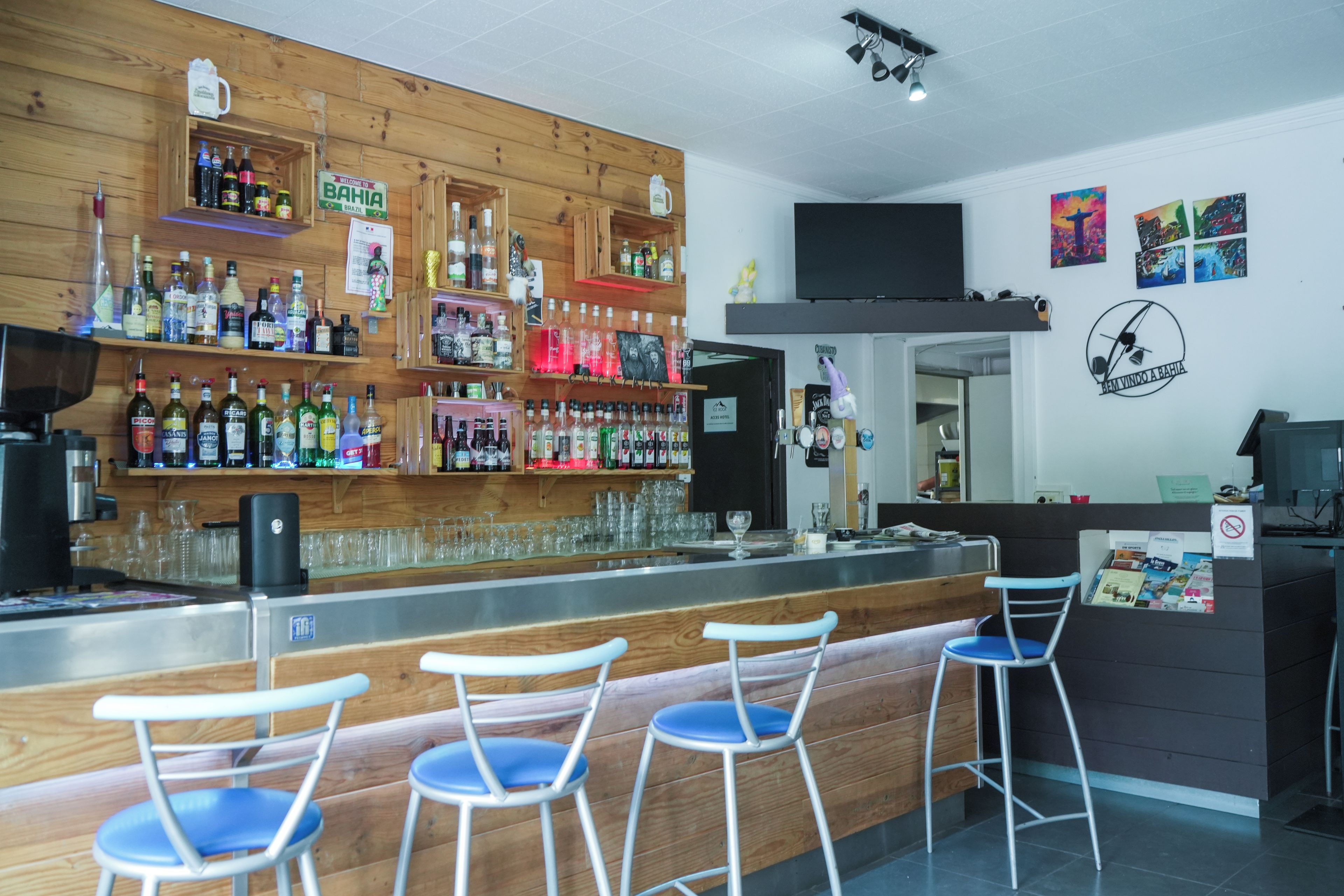 A bar area with wooden shelves stocked with various liquor bottles, a flat-screen TV mounted on the wall, and a bar counter with stools.