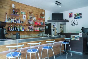 A bar area with wooden shelves stocked with various liquor bottles, a flat-screen TV mounted on the wall, and a bar counter with stools. - Ice Rock Brasil - Hôtel et restaurant Brésilien (L'Argentière-la-Bessée)