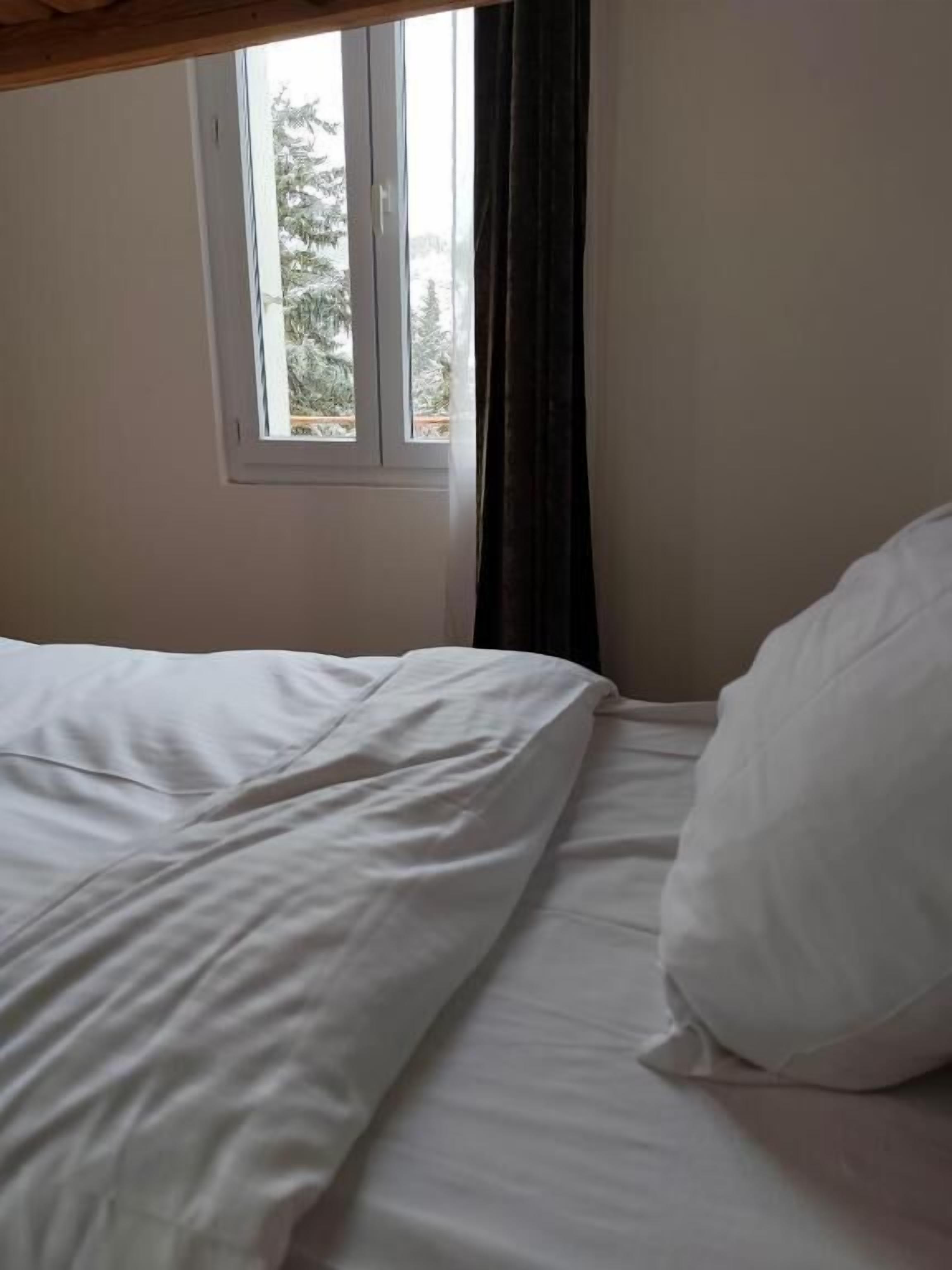 Una cama con ropa de cama blanca, una ventana con vistas a árboles y una cortina oscura.