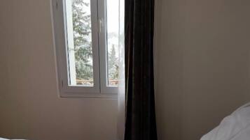 Una cama con ropa de cama blanca, una ventana con vistas a árboles y una cortina oscura.
