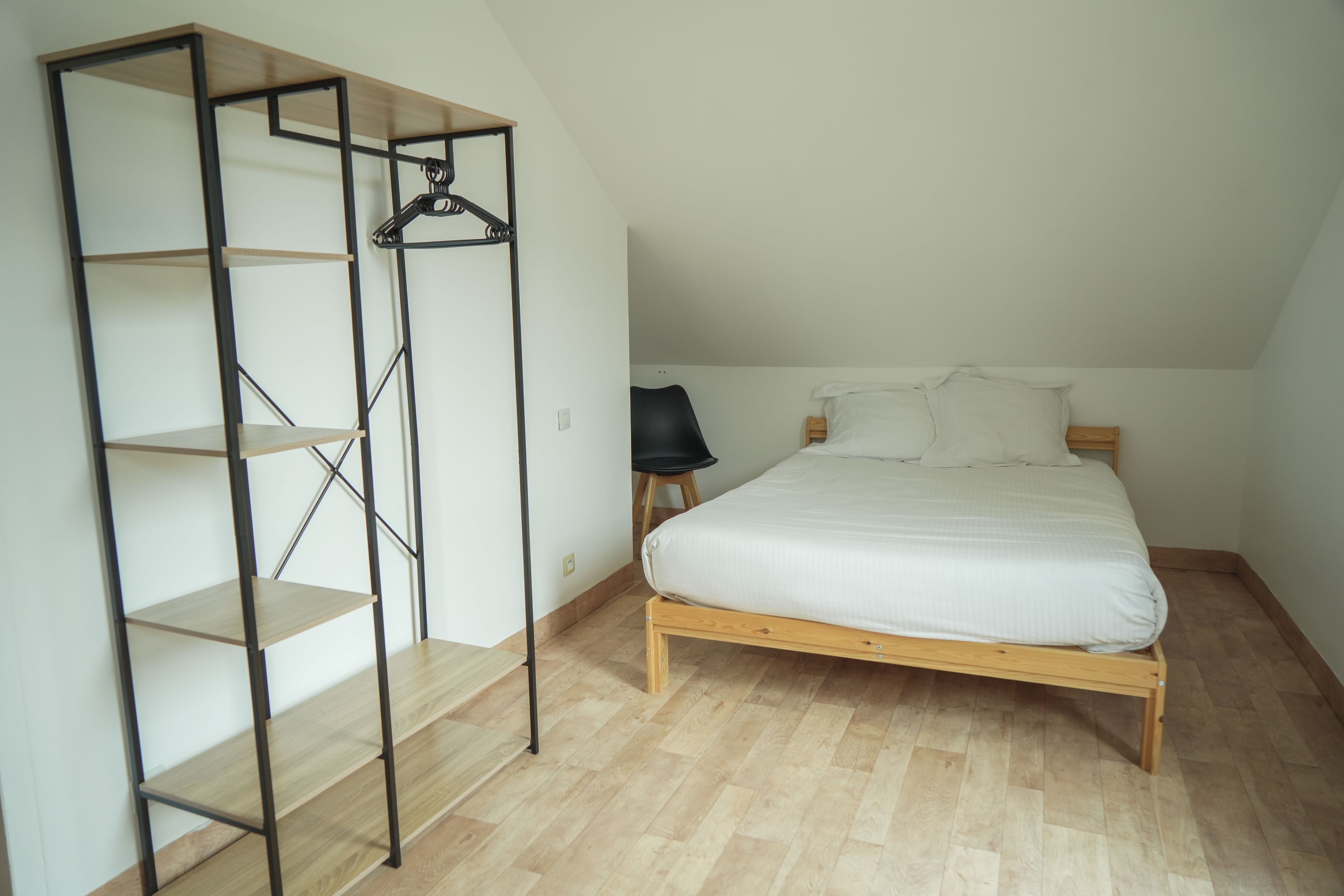 Un dormitorio con una cama de madera, una silla negra y un estante metálico.