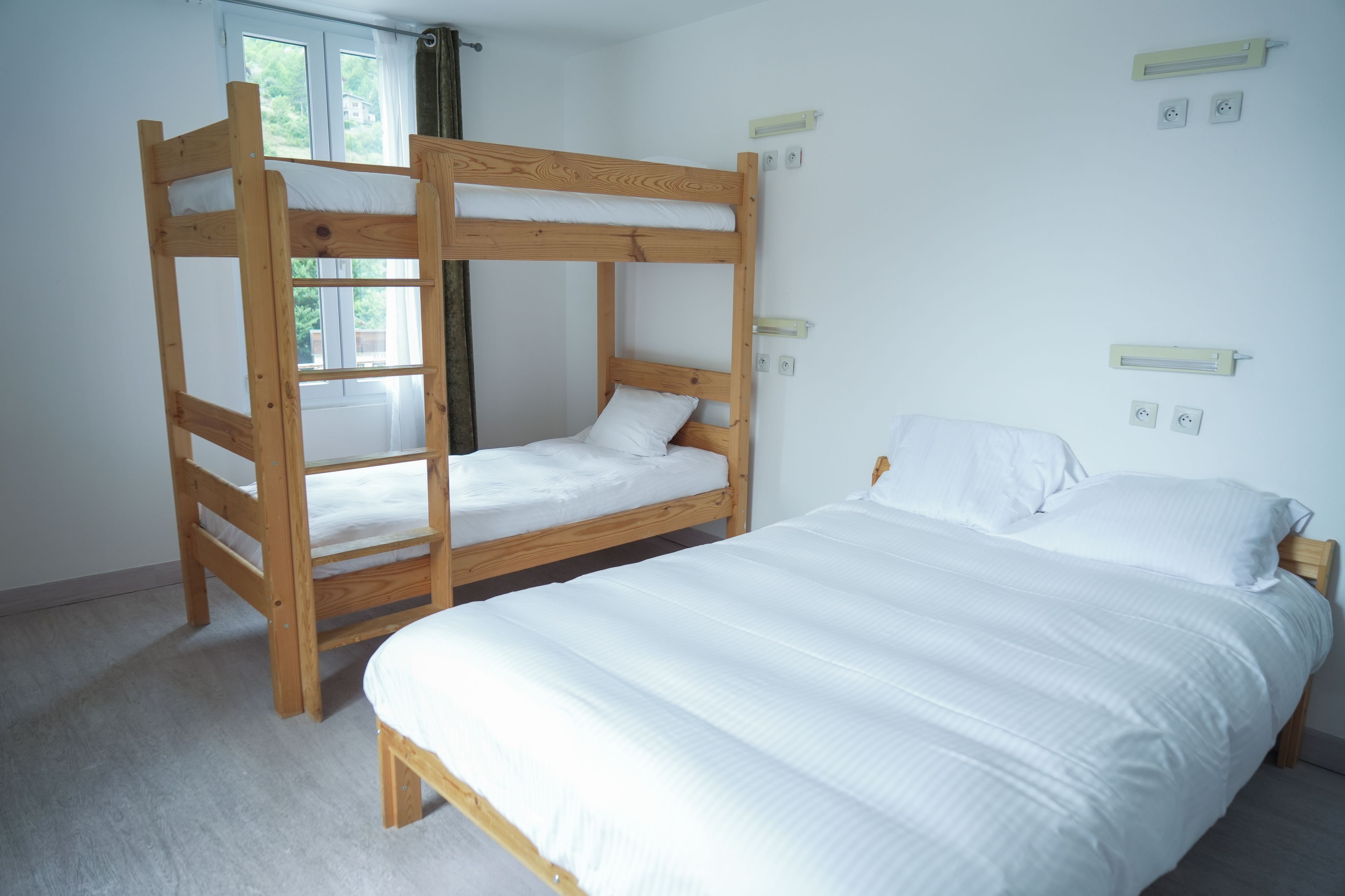 A bunk bed with a lower bunk and a single bed, both with white bedding. There are two wall-mounted light switches and a window with curtains.