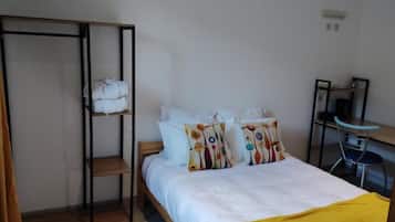 Una habitación con una cama, un estante con ropa doblada, un escritorio con silla y un espejo.