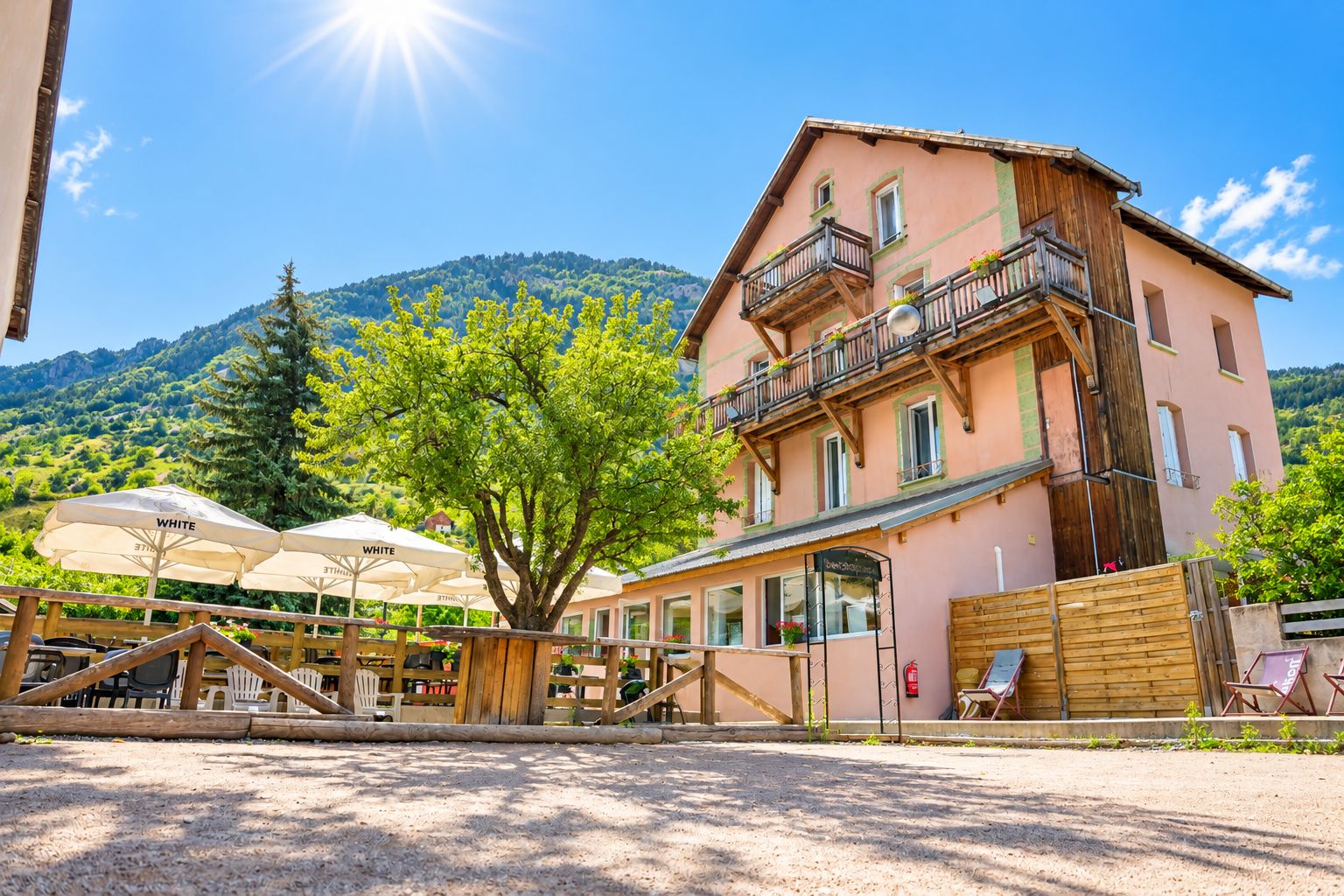 Un edificio a due piani con un balcone, un'area salotto all'aperto con ombrelloni e un paesaggio montuoso sullo sfondo.