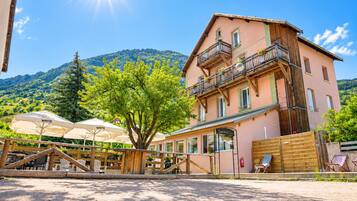 Un edificio a due piani con un balcone, un'area salotto all'aperto con ombrelloni e un paesaggio montuoso sullo sfondo.