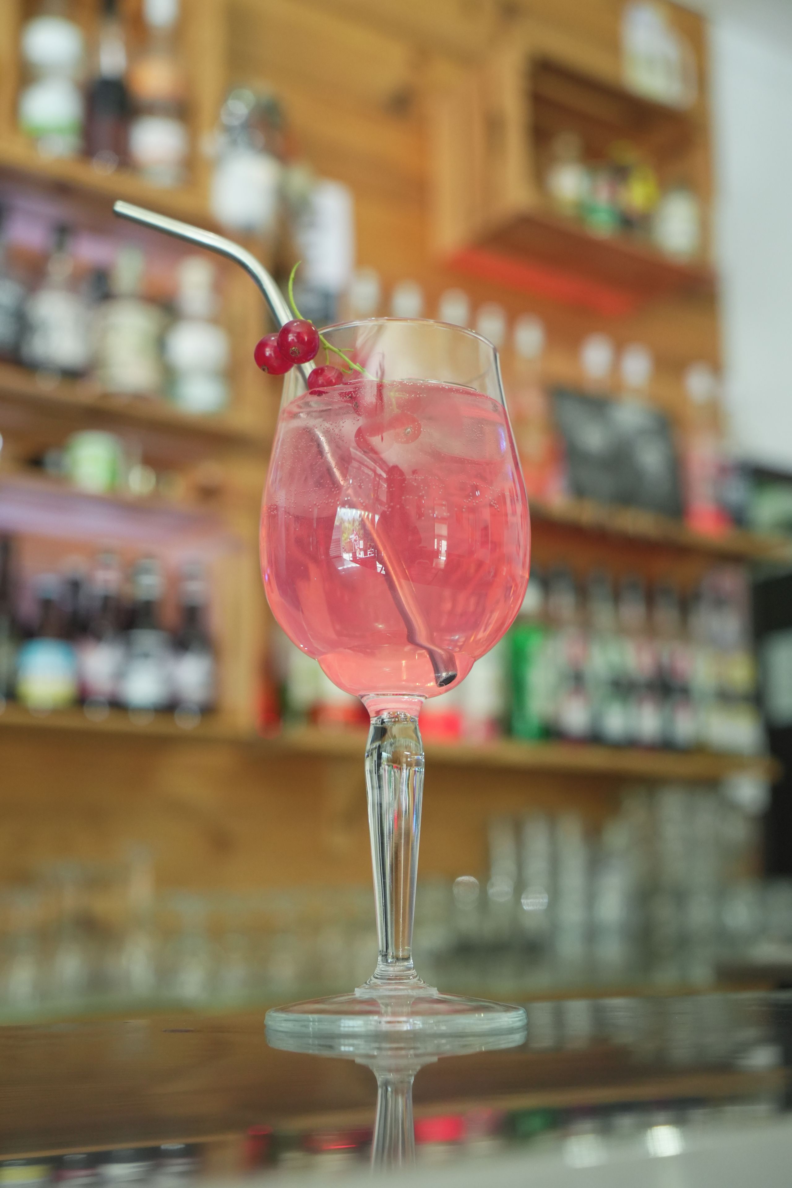 A glass with a pink beverage, garnished with a cherry and a straw, on a bar counter with shelves in the background.