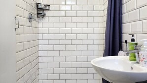 A shower with white tiles, a showerhead, a dark curtain, and a sink with toiletries.
