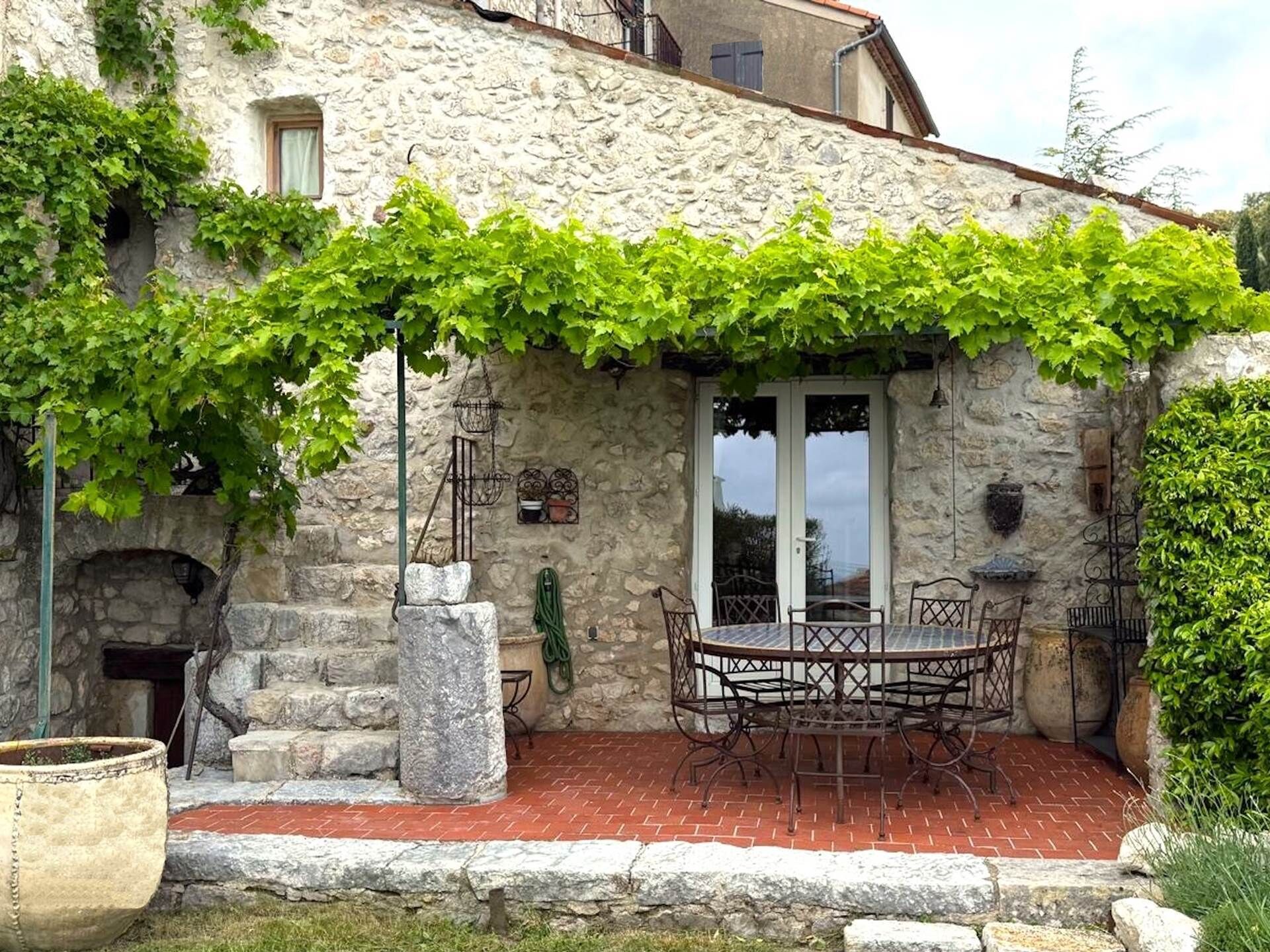 A stone building with a vine-covered pergola, a brick patio, and a table with chairs.