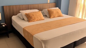 Una cama bien hecha con cabecera de madera, una mesita de noche con lámpara, y otra mesita con un teléfono y un libro.