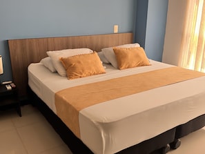 Una cama bien hecha con cabecera de madera, una mesita de noche con l ámpara, y otra mesita con un teléfono y un libro.