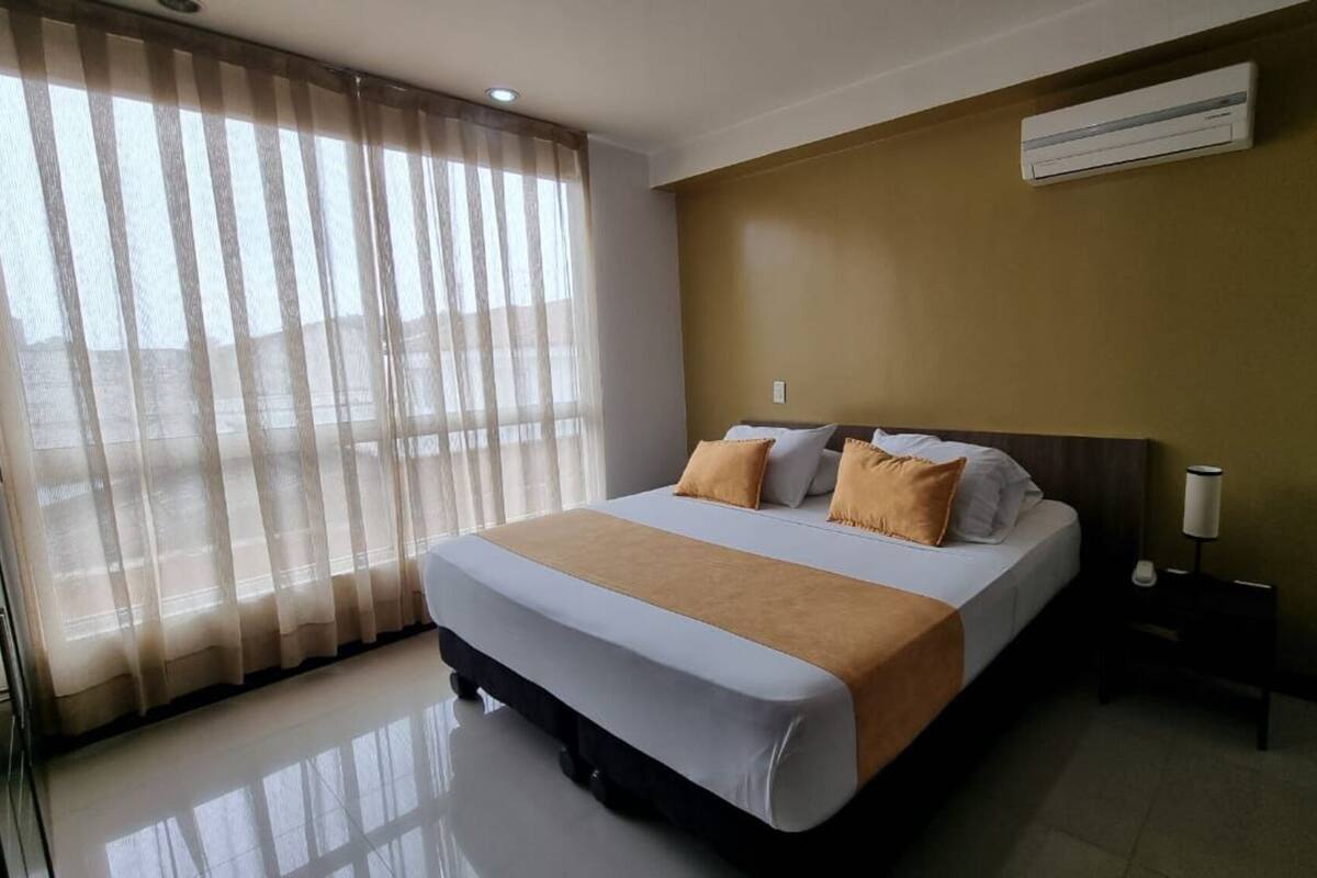 Une chambre d’hôtel avec un lit, une table de chevet, une lampe et une fenêtre avec des rideaux légers.
