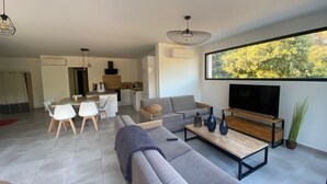 Una sala de estar moderna con sofá, mesa de centro y televisor. Se observa un comedor con sillas y una cocina al fondo.