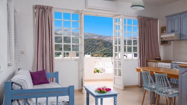 A room with a bed, a table with flowers, a kitchen area, and a balcony with a mountain view. - Apartment 'Memories' with Balcony, Wi-Fi and Air Conditioning (Palekastro)