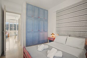 A bedroom with a bed, a nightstand, a lamp, a wardrobe, and a view through an open door. - Apartment 'Memories' with Balcony, Wi-Fi and Air Conditioning (Palekastro)
