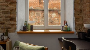 Una habitación con pared de ladrillo, ventana con vista a otro edificio de ladrillo, una silla de madera, una mesa y una estantería con diversos objetos.