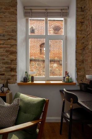 Ein Zimmer mit Backsteinwand, einem Fenster mit Blick auf ein Backsteingebäude, einem Holzstuhl, einem Tisch und einem Regal mit verschiedenen Gegenständen.