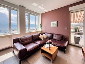 Ein modernes Wohnzimmer mit einer braunen Ledersofa, einem Holztisch und großen Fenstern mit Blick auf das Meer und die Stadt.