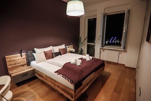 Une chambre à coucher avec un lit en bois, une table de chevet, une chaise, une fenêtre donnant sur les lumières de la ville et un luminaire suspendu.