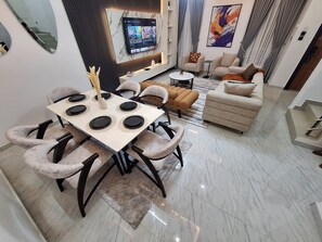 A modern living room with a dining area, a flat-screen TV mounted on the wall, and a sofa set. - Spacious 4-bedroom house with 24hr-Power Supply & Security, Gym, Pool (Lekki)