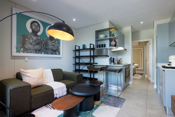 A modern living room with a sofa, coffee table, and bar stools. There is a kitchen area with a bar counter and shelves. A large painting is on the wall. - Slick Garden Gem 300 on Florida RD (Morningside, KwaZulu-Natal)