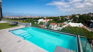 Una piscina en la azotea con vista a la ciudad, un área con césped y una tumbona.