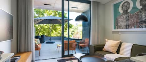 A modern living room with a sofa, coffee table, and chairs, a sliding glass door leading to an outdoor patio with umbrellas and seating.