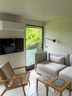 Un salon moderne comprenant un canapé, une chaise en bois, une petite table, un téléviseur fixé au mur et une vue sur la verdure grùce à une porte coulissante.